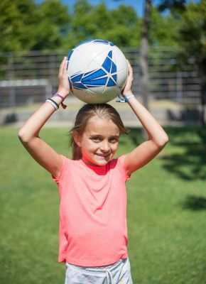 pexels photo 8813514 8813514 Smiling girl holding a soccer ball above her head on a sunny day outdoors.